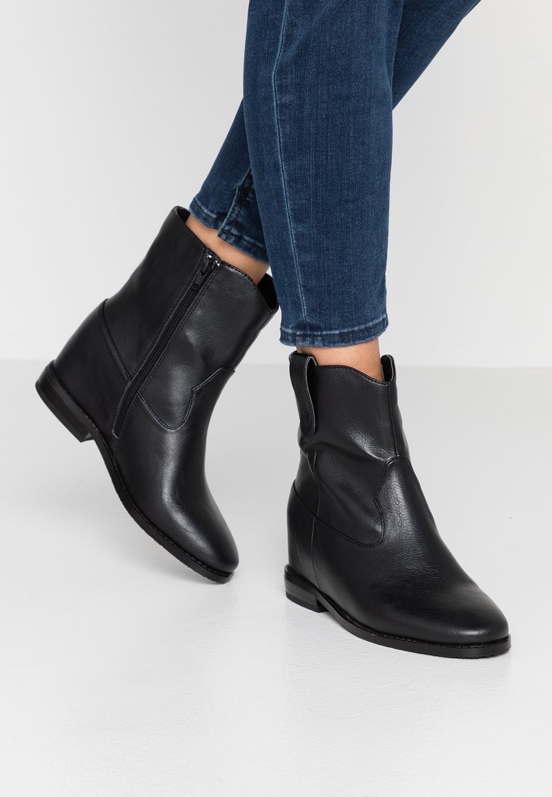 Bottes noires en cuir à la cheville avec fermetures éclair latérales portées avec un jean skinny bleu foncé retroussé sur un sol blanc.