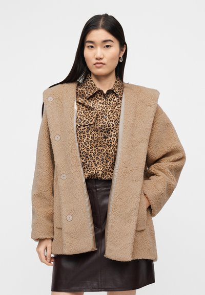 Μπεζ, χνουδωτό παλτό με υπερμεγέθη γιακά και κουμπιά στο κλείσιμο, φορεμένο πάνω από καφέ πουκάμισο με λεοπάρ λεκέδες και σκούρα δερμάτινη φούστα.