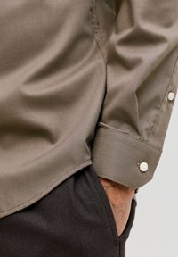 Καφέ χακί πουκάμισο με υφή, με μυτερό γιακά και αντίθετα λευκά κουμπιά, συνδυασμένο με σκούρο παντελόνι.