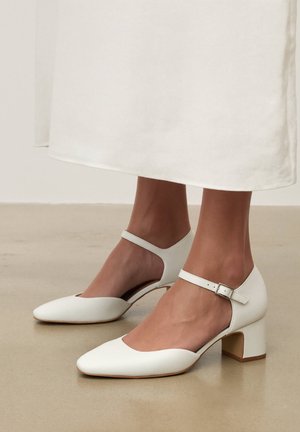 Woman wearing white leather Mary Jane heels with block heels and ankle straps, standing on beige floor with white skirt hem visible.