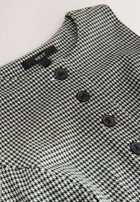 Tessuto a motivo pied de poule in bianco e nero con finitura testurizzata. Presenta un colletto e tre bottoni scuri sul davanti.