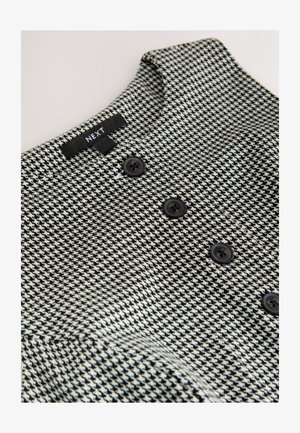 Stof met een houndstooth-patroon in zwart-wit en een gestructureerde afwerking. Voorzien van een kraag en drie donkere knopen aan de voorkant.