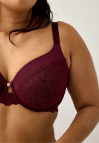 Burgundifärgad bh med spetsdetaljer och justerbara remmar, med en rosa knappaccent i mitten, slät tyg och vågiga kanter.