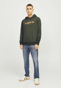 Sudadera verde con logo naranja, cordones y puños acanalados, combinada con jeans azules y zapatillas blancas con acentos verdes y rojos.