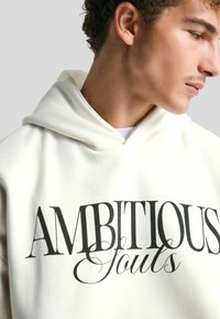 Fehér kapucnis pulóver, amelynek elején merész fekete felirat olvasható: "AMBICIOZUS Lelkek". Puha, sima textúrával és laza szabású dizájnnal rendelkezik.