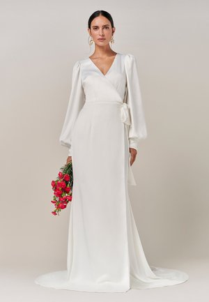 Robe blanche à manches bouffantes longues, avec un détail de taille nouée et une texture satinée lisse, portée avec un bouquet de fleurs roses.