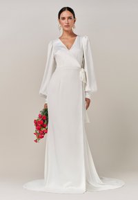 Weißes Wickelkleid mit langen Puffärmeln, das über eine gebundene Taillendetail und eine glatte Satinoberfläche verfügt, getragen mit einem Strauß pinkfarbener Blumen.