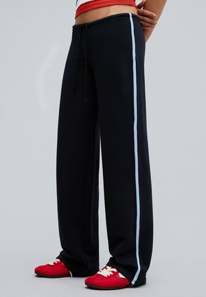 Personne portant un pantalon noir à jambes larges avec des bandes latérales bleu clair et des baskets rouges à lacets blancs, debout les mains derrière le dos.