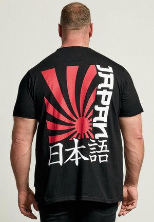 Mand iført sort T-shirt med rød opadgående solgrafik og hvid lodret japansk tekst på bagsiden, stående foran en ensfarvet baggrund.