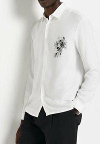 Camicia bianca a maniche lunghe con stampa floreale nera e grigia sul lato sinistro del petto, caratterizzata da un colletto classico e chiusura con bottoni.