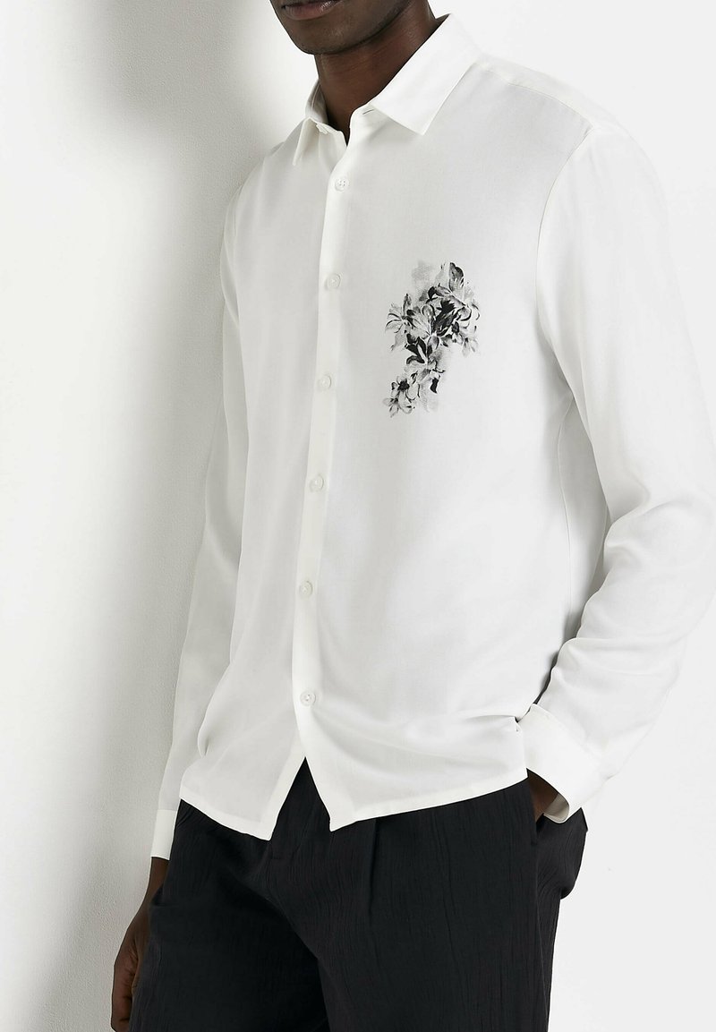 Camicia bianca a maniche lunghe con stampa floreale nera e grigia sul lato sinistro del petto, caratterizzata da un colletto classico e chiusura con bottoni.