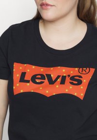 Gros plan d'une personne portant un t-shirt Levi's noir avec un logo orange orné de petits motifs floraux jaunes.