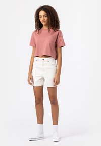 T-shirt rosa tagliato con tasca frontale, abbinato a pantaloni shorts in denim bianchi. Il modello indossa sneakers bianche e calze bianche fino al ginocchio.