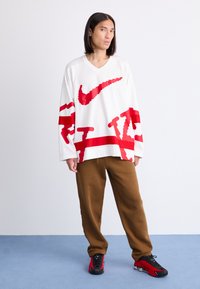 Hvid langærmet mesh Nike top med røde grafikker og V-udskæring. Brune sweatpants og rød/sorte sko. Simpel baggrund, stående positur.