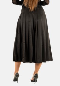 Femme aux cheveux longs portant une jupe midi plissée noire et des chaussures à talons hauts noires et brillantes, se tenant sur un fond blanc.