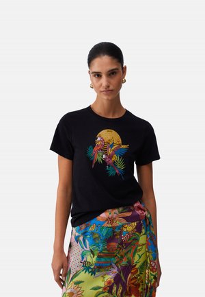 Donna che indossa una maglietta nera con un design colorato di pappagalli e sole, abbinata a una gonna vivace con motivi floreali e fogliari.