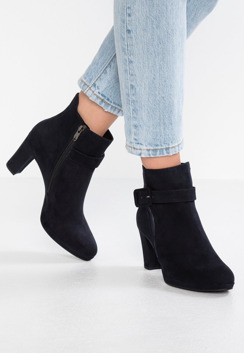 Femme portant des bottines en daim noir à talons blocs et fermeture éclair latérale, associées à un jean court bleu clair.