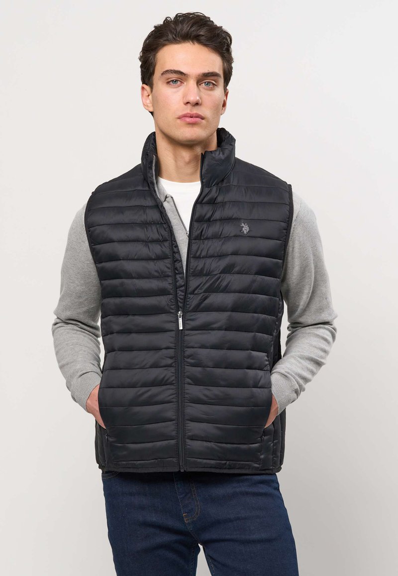 Mladić s tamnom kosom nosi crni prošiveni prsluk preko sive majice i plave traperice, stojeći pred jednostavnom svijetlom pozadinom.