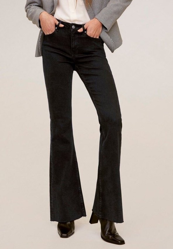Femme portant un jean flare taille haute noir, des bottes pointues noires, un chemisier blanc et un blazer gris avec les mains dans les poches.