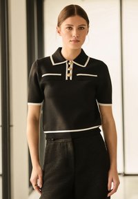 Schwarzes gestricktes Poloshirt mit kurzen Ärmeln, weißen Akzenten und drei dekorativen Knöpfen. Verfügt über zwei kleine Brusttaschen.