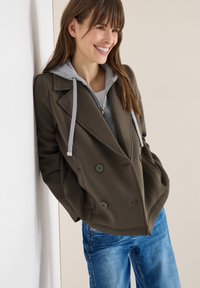 Femme souriante, portant un manteau croisé vert olive foncé par-dessus un sweat à capuche gris, appuyée contre un mur blanc, avec un jean bleu.