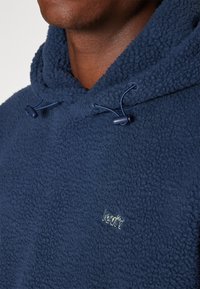 Felpa con cappuccio in pile blu navy con superficie testurizzata, coulisse regolabile e logo ricamato discreto sul petto. Materiale morbido e caldo.