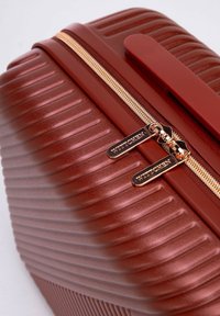 Valigia rigida rossa con texture, caratterizzata da scanalature verticali, chiusura con zip dorata e hardware inciso. Forma compatta con una finitura liscia.