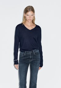 Námořnický modrý svetr ve výstřihu do V vyrobený z měkké tkaniny, spárovaný s tmavými džínovými kalhotami s viditelným prošíváním a předními kapsami.