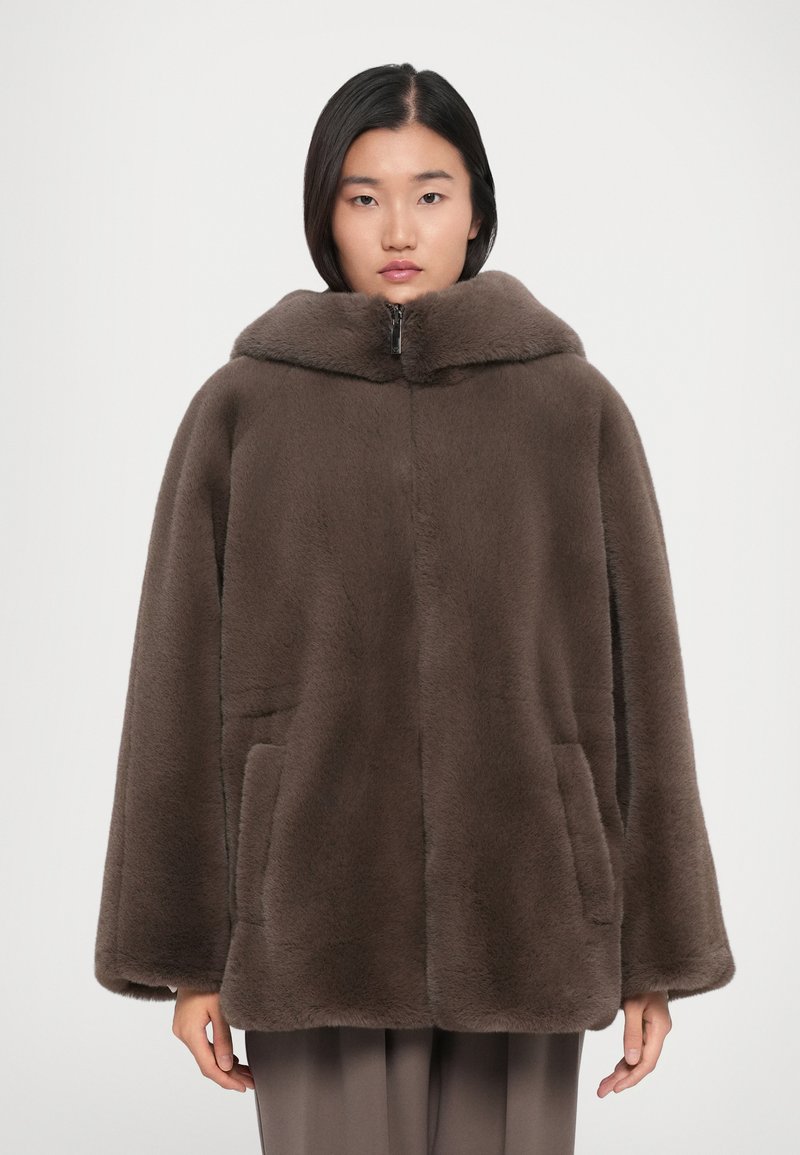 Kvinna i en brun zip-up jacka i konstfuskpäls med en bred krage och framfickor, står mot en enkel ljus bakgrund.