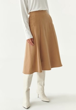 Femme portant une jupe midi évasée couleur camel avec des poches latérales et des bottes blanches à talons montant jusqu'aux genoux, debout devant un fond neutre.
