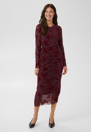Femme aux longs cheveux bruns portant une robe midi fleurie rouge foncé avec de longues manches transparentes et des talons noirs, souriante devant un fond uni.