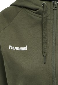Hoodie zippé vert olive en tissu doux, présentant un petit logo blanc "hummel", un cordon texturé et des accents côtelés.
