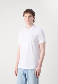 Young man with short brown hair wearing a white polo shirt and light blue jeans standing against a plain background.