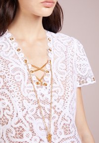 Kobieta w białej koronkowej bluzce z sznurowaniem z złotych łańcuchów i oczkami, ukazująca szczegółowe kwiatowe i geometryczne wzory koronki na rękawach i klatce piersiowej.