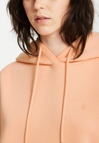 Hoodie w kolorze brzoskwiniowym z sznurkiem, wykonany z miękkiego materiału. Posiada małe logo na klatce piersiowej oraz klasyczny design kieszeni kangurkowej.
