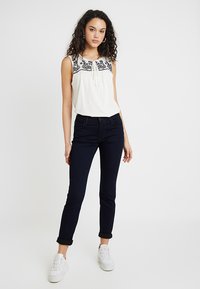 Blusa blanca sin mangas con bordado floral negro y detalle de lazo en el escote, combinada con jeans azul oscuro y zapatillas blancas.