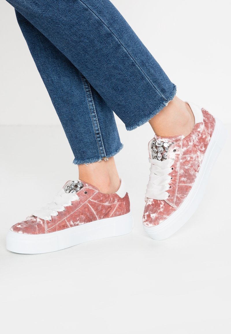 Baskets en velours rose avec une semelle en caoutchouc blanche, dotées d'accents métalliques sur la languette et de lacets plats blancs. Pantalon court en denim également visible.