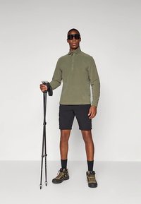 Man i solglasögon som bär en grön zip-up-tröja, svarta shorts, vandringskängor, håller två vandringsstavar mot en enkel grå bakgrund.