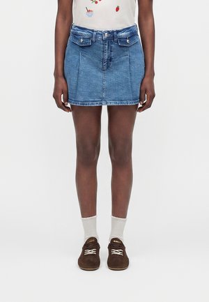 Personne portant une mini-jupe en jean bleu avec des poches à rabat à l'avant, des chaussettes blanches, des baskets marron et une chemise blanche avec des motifs de fruits.