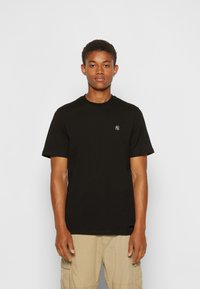 Black cotton t-shirt with round neck and short sleeves, featuring a small white logo on the chest. Worn with beige cargo shorts.