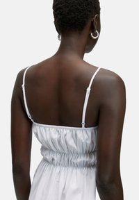 Weißes Kleid mit dünnen Trägern, gerafftem Rücken, glatter Textur und ausgestelltem Schnitt, vor einem neutralen Hintergrund.