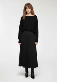 Maglione nero oversize con scollo ampio, abbinato a una gonna midi plissettata nera. La modella indossa stivaletti neri con tacco. Texture liscia.