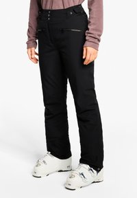 Schwarze Skihosen mit hoher Taille, ausgestattet mit Reißverschlusstaschen, Knopfverschluss und einer glatten Textur, kombiniert mit weißen Skischuhen.