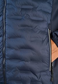 Mörkblå vadderad jacka med quiltad textur, utrustad med sidodragkedja och slät tygkvalitet. Utsmyckad med en kontrasterande meshnätspanel.