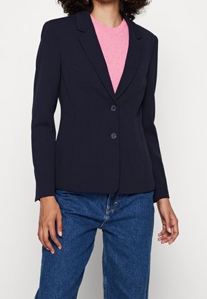 Marineblazer mit taillierter Passform, scharfen Revers, Zwei-Knopf-Verschluss und glatter Textur; getragen über einem rosa Pullover, kombiniert mit blauen Jeans.