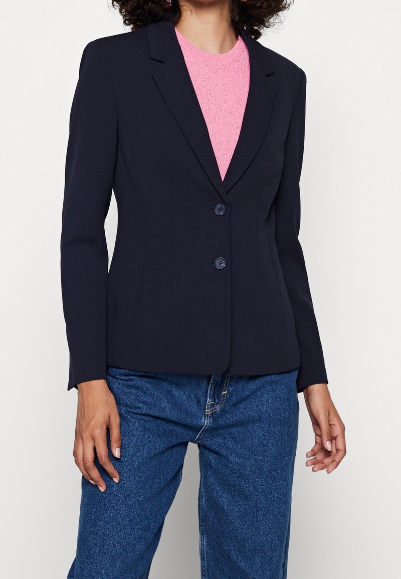 Marinblå blazer med skräddarsydd passform, skarpa slag, tvåknappsstängning och mjuk textur; bärs över en rosa tröja, tillsammans med blå jeans.