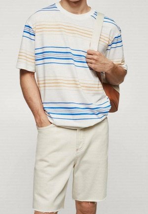 Hombre con pantalones cortos blancos y camiseta blanca con rayas azules y beige, sosteniendo la correa de un bolso bandolera marrón claro y blanco.