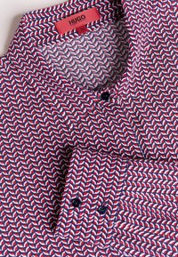 Camicia a maniche corte con un motivo geometrico blu navy, rosso e bianco. Presenta una patta con bottoni e un colletto a ribaltina. Realizzata in tessuto.