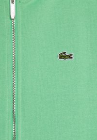 Sweat à zip vert avec un design en tricot texturé et un logo crocodile Lacoste brodé sur la poitrine gauche. Quincaillerie de zip en argent visible.
