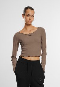 Pruun ribadega pikad varrukad crop top, millel on sakiline äär ja logo tekst "KANI" ees, koos mustade lõdva lõikega pükstega.